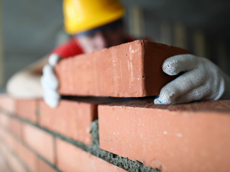Close-up of professional construction worker laying bricks in industrial site. Builder in protective uniform. Man building wall with blocks. Renovation concept